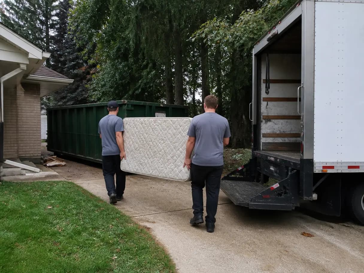 A junk removal crew carrying a mattress past a roll-off in a driveway.