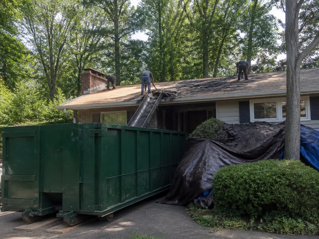 A roofing crew tearing shingles off a house into a dumpster in the driveway.